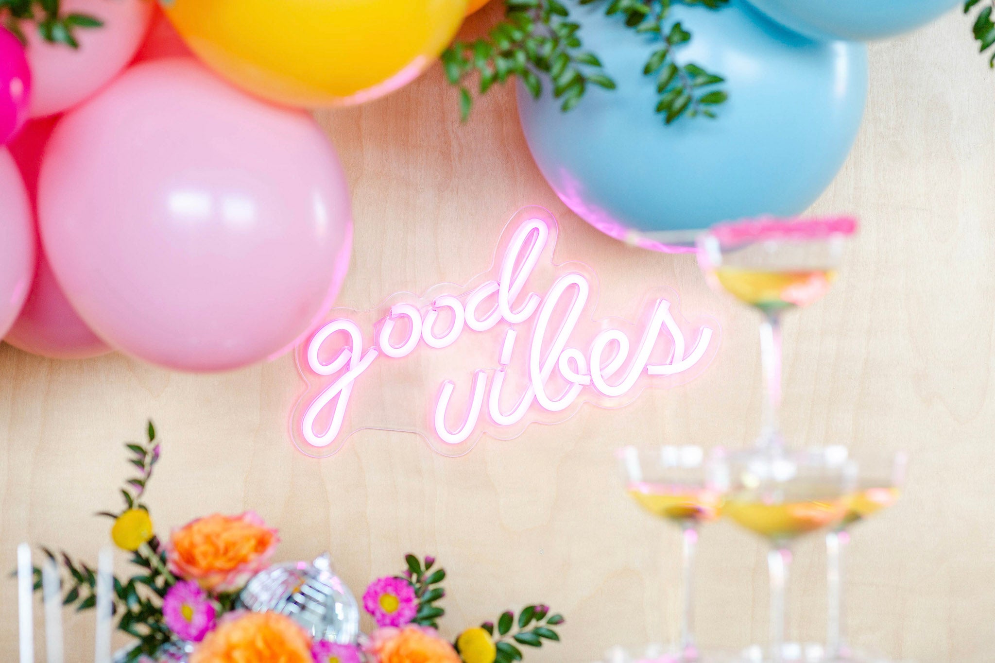 Neon sign saying 'good vibes' with balloons and flowers on a wooden surface